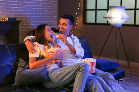 Happy young couple with popcorn watching movie at home in the eveningの写真素材