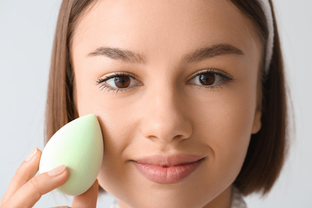 Pretty young woman with green makeup sponge on gray background, closeupの写真素材