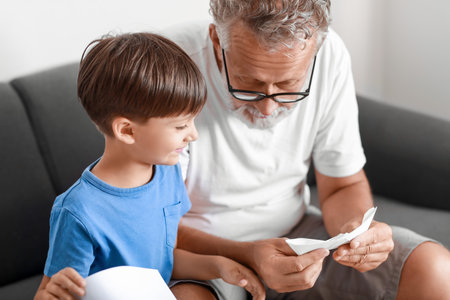 Senior man showing his little grandson how to make paper boat at homeの写真素材