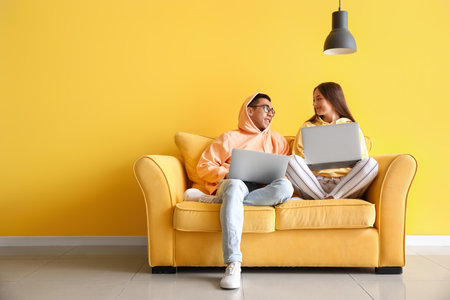 Young couple with laptops sitting on sofa near yellow wallの写真素材
