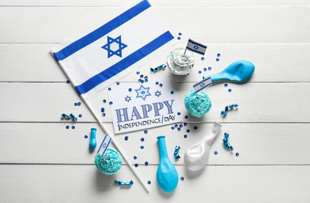 Card with text HAPPY INDEPENDENCE DAY, flag of Israel, cupcakes and festive decor on white wooden backgroundの写真素材