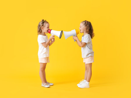 Cute little girls shouting into megaphones on yellow backgroundの写真素材