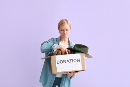 Young woman holding box with donation clothes on lilac backgroundの写真素材