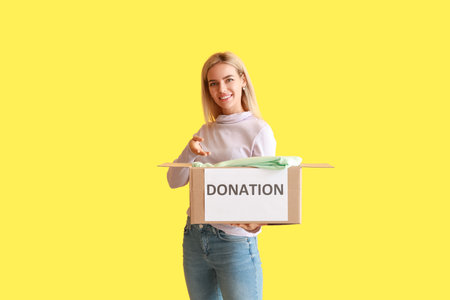 Young woman holding box with donation clothes on yellow backgroundの写真素材