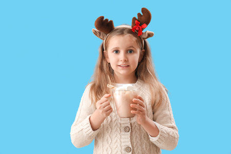 Little girl in reindeer horns holding cup of cocoa with marshmallows on blue backgroundの写真素材