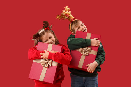 Little children in reindeer horns hugging Christmas presents on red backgroundの写真素材
