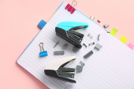 Open notebook, staplers and staples on pink background