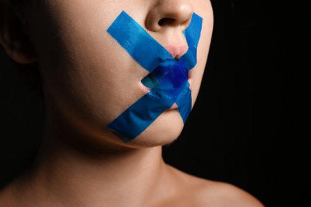 Young woman with taped mouth on dark background, closeup. Censorship conceptの写真素材