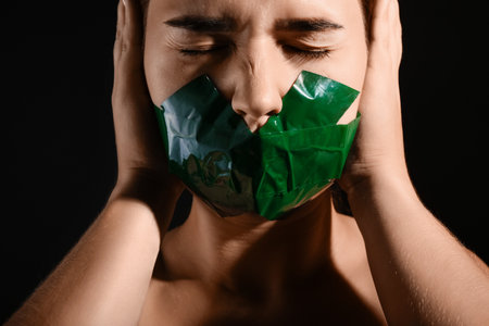 Young woman with taped mouth covering ears on dark background. Censorship concept