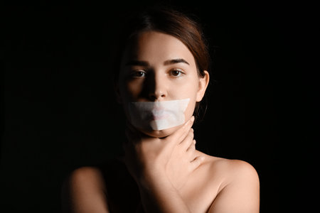 Young woman with taped mouth on dark background. Censorship conceptの写真素材