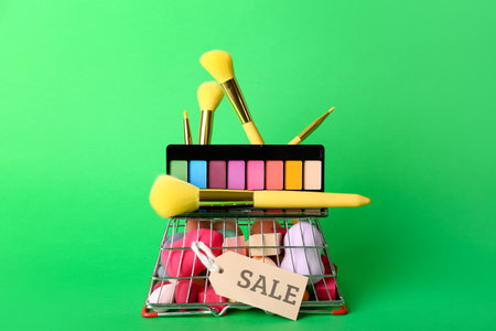 Shopping basket with makeup brushes, sponges, eyeshadows and sale tag on green backgroundの写真素材