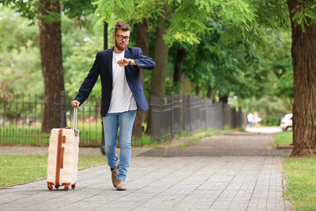 Handsome young man with suitcase looking at wristwatch outdoorsの写真素材
