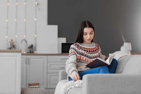 Young woman reading book on sofa in kitchenの写真素材