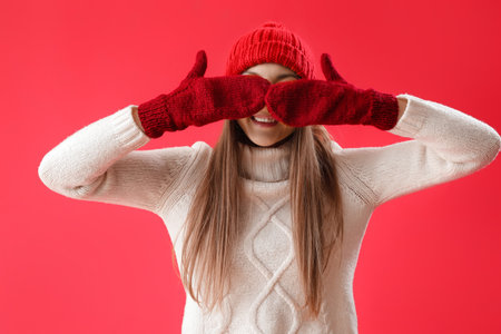 Young woman in warm hat and mittens closing eyes on red backgroundの写真素材