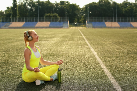 Young woman with headphones meditating on grass at sport stadiumの写真素材