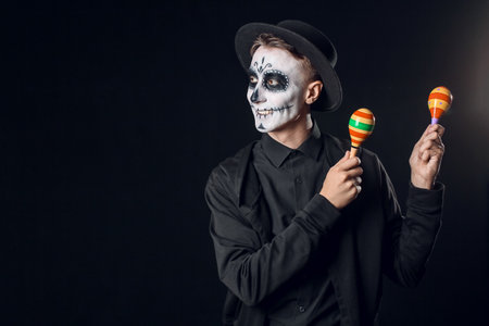 Young man with painted skull on his face and maracas against dark background. Celebration of...