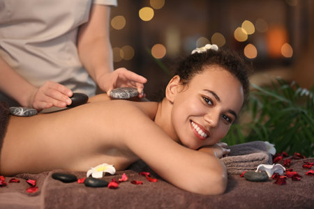 Relaxed young African-American woman getting massage with stones by therapist in spa salonの写真素材