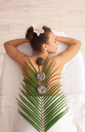 Young African-American woman with palm leaf and stones lying on couch in spa salon