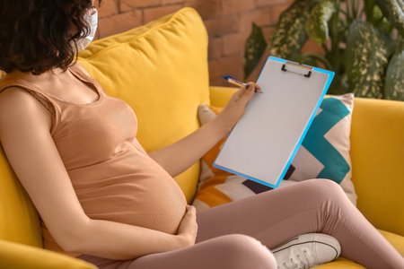 Young pregnant woman in medical mask with clipboard at homeの写真素材