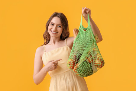 Young woman holding string bag with food on yellow backgroundの写真素材