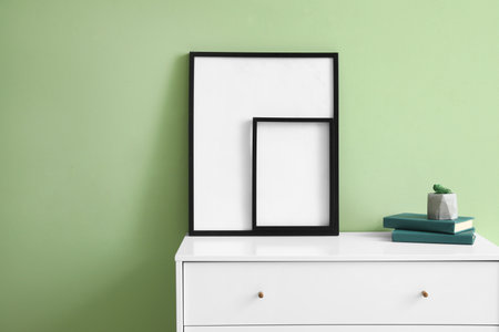 Blank frames and books on chest of drawers near green wallの写真素材
