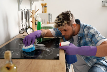 Handsome young man with sponge cleaning stove in kitchenの写真素材