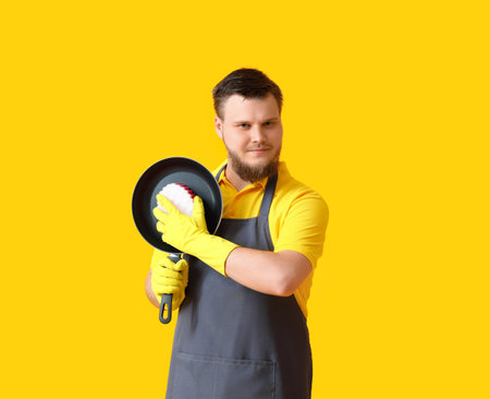 Young man with sponge cleaning frying pan on yellow backgroundの写真素材