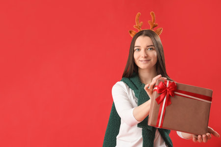 Young woman in reindeer horns with Christmas gift on red backgroundの写真素材