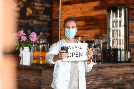 Male business owner with medical mask, coffee cup and opening sign in his cafe