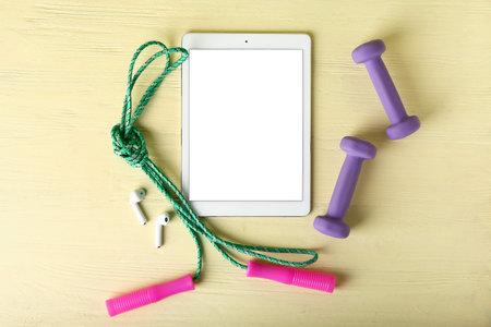 Tablet computer with earphones, skipping rope and dumbbells on yellow wooden background