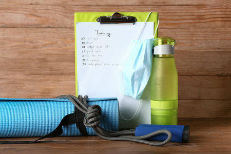 Clipboard with training schedule, medical mask, mat, bottle of water and skipping rope on wooden...