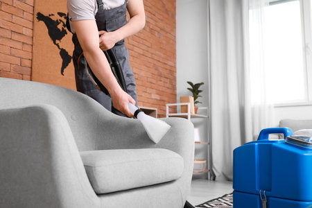 Male worker removing dirt from gray armchair with vacuum cleaner in roomの写真素材