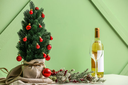 Small Christmas tree with glass and bottle of wine on table near green wallの写真素材