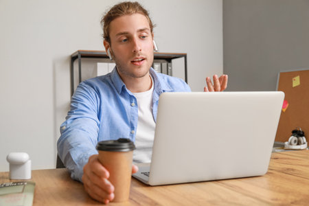 Handsome young man with cup of coffee video chatting in office