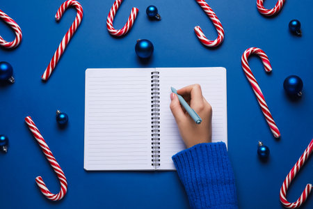 Female hand with notebook, pen, candy canes and Christmas balls on color backgroundの写真素材