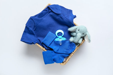 Basket with baby clothes, pacifier and toy on light background