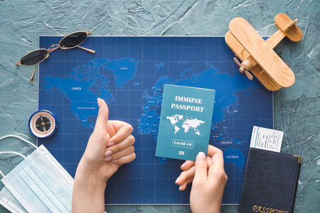 Hands of tourist with travel supplies and immune passport showing thumb-up on color background