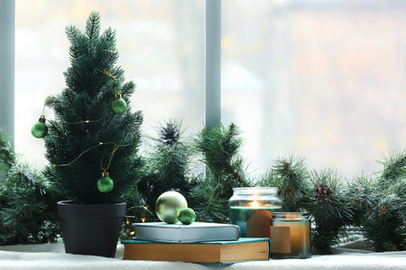 Small Christmas tree with balls, books, candles and fir branches on windowsill in roomの写真素材