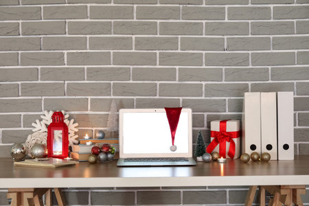 Laptop with Santa hat, Christmas decor and presents on table near gray brick wallの写真素材