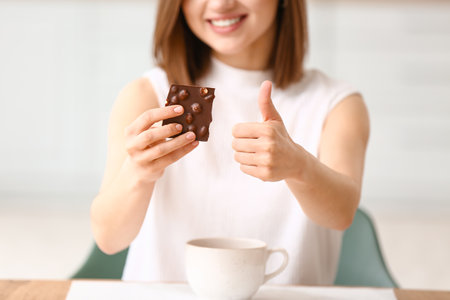 Beautiful young woman with tasty chocolate showing thumb-up at homeの写真素材