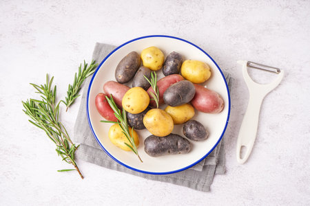 Plate with different raw potatoes, rosemary and peeler on light backgroundの写真素材