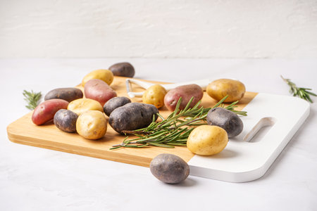 Wooden board with different raw potatoes and rosemary on light backgroundの写真素材