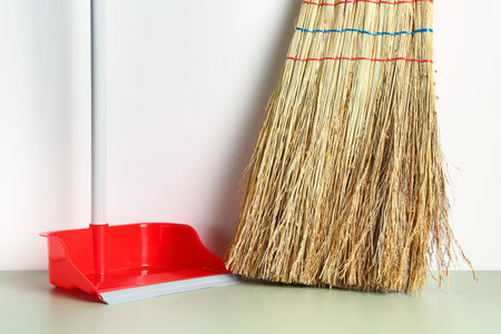 Red dustpan and broom near light wall, closeup