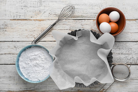 Baking dish with paper, whisk, bowls, flour and eggs on white wooden backgroundの写真素材