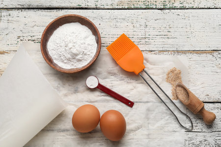 Baking paper with kitchen supplies, bowl of flour and eggs on white wooden backgroundの写真素材