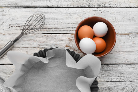 Baking dish with paper, whisk, bowl and eggs on white wooden background