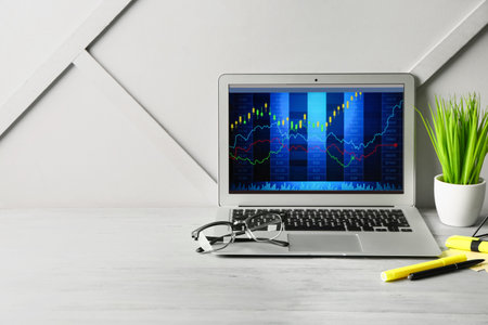 Laptop with trading charts and eyeglasses on table near light wall