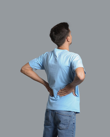 Young man suffering from back pain on gray background