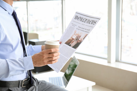 Young businessman with cup of coffee reading newspaper in officeの写真素材