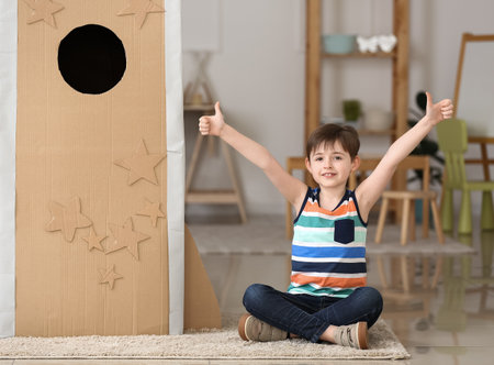 Cute little boy with cardboard rocket showing thumb-up at homeの写真素材